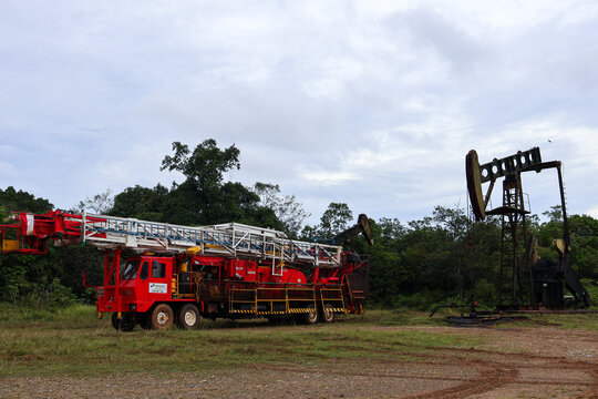 Tanjung-Indonesia May 17, 2026 A heavy-duty red mobile well servicing workover rig truck parked next to an operating oil pumpjack unit at an extraction site.