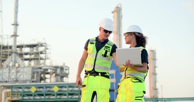 Male technician discusses refinery workflow while female engineer reviews laptop data beside petrochemical structure during industrial maintenance inspection and planning session