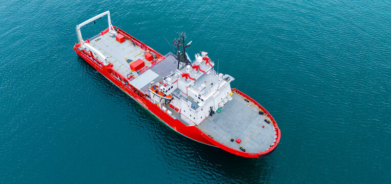 Cinematic aerial drone perspective showcasing the aft deck and command bridge of a heavy industrial red and white ship cruising through calm ocean waters during a commercial voyage.