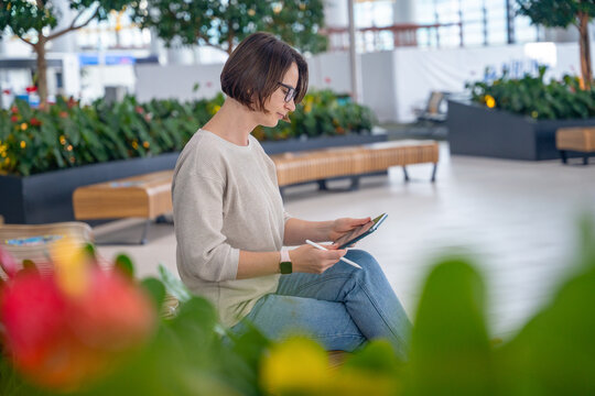 Young woman with glasses in beige sweater sitting on bench using tablet in bright modern indoor garden atrium with colorful flower in foreground, lifestyle remote work concept.