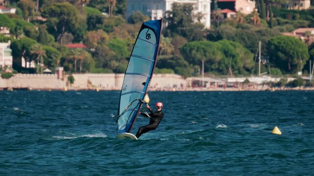 Windsurfer gliding fast across blue sea surface near coastal town on sunny day with dynamic motion.