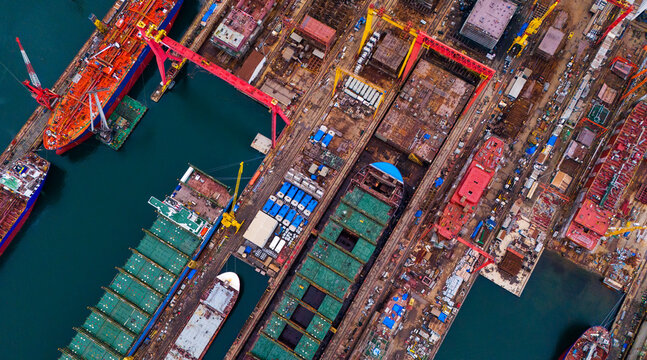 Top down aerial shot of active shipbuilding facility showcasing giant red gantry crane over dry dock large green cargo ship holds and various marine manufacturing tools