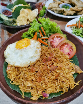 Indonesian Mie Goreng. Savory fried noodles served on a banana leaf in a rustic clay plate, topped with a sunny-side-up fried egg, crispy shallots, sliced tomatoes, and fresh vegetables