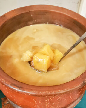 Close-up view of a traditional Indonesian sweet dessert, Kolak Ketela, served warm in a rustic earthenware clay pot. A spoon is lifting chunks of sweet potato cooked in a rich broth of coconut milk