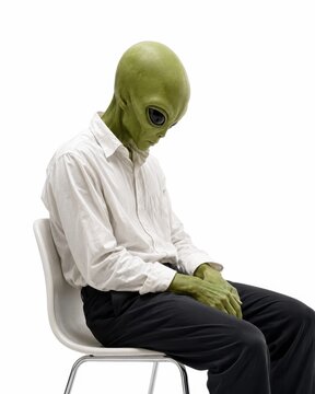 Sad green alien quiet office burnout concept loose white shirt and simple dark trousers sitting alone on a minimalist chair head gently lowered hands resting on lap