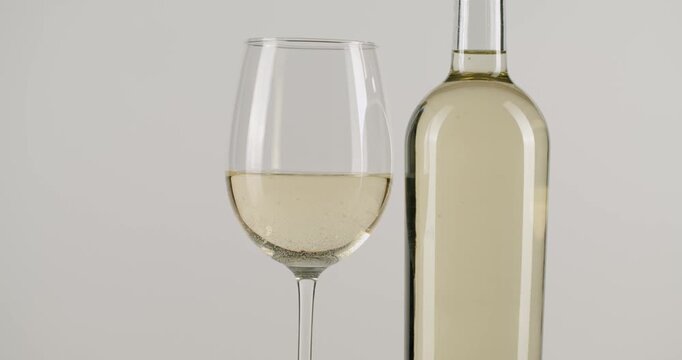 Wineglass full of chilled white wine swirling next to a bottle on a white background, with small bubbles gracefully rising in the golden liquid in a beautiful slow motion shot.