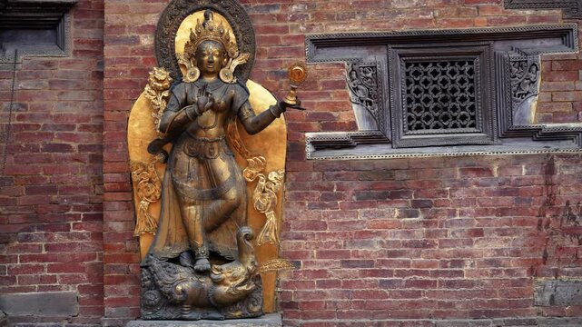 View of a golden deity statue standing against a traditional red brick wall with ornate wooden carvings in Nepal.