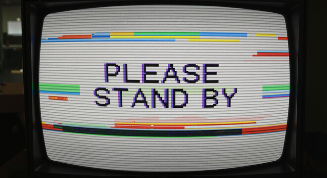 Retro television screen displaying 'PLEASE STAND BY' message with colorful glitchy lines symbolizing technical difficulties or broadcast interruption.