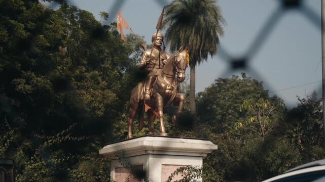 Gilded equestrian monument of the Maratha warrior king Chhatrapati Shivaji Maharaj seen through a fence, standing among lush green trees under a clear blue sky in India