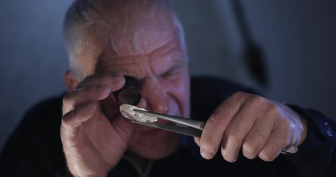 Closeup tracking shot of elderly jeweler inspecting rough diamond with loupe and tweezers at dim indoor workshop bench.