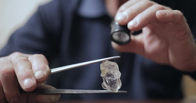 Closeup jeweler examining rough diamond on workbench with loupe and tweezers.