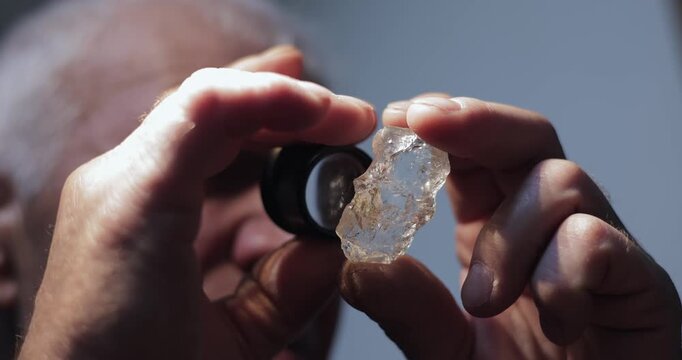 Handheld close up man examining mineral diamond gemstone with loupe and rotating clear crystal against neutral studio background.