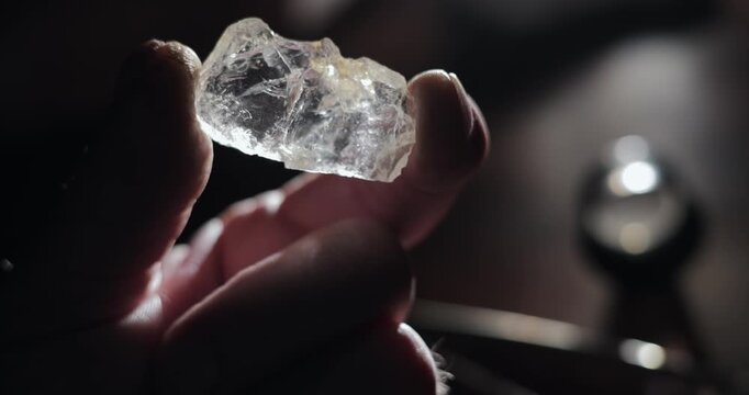 Closeup slow motion hand holding rough diamond crystal in dark workshop on backlit table.