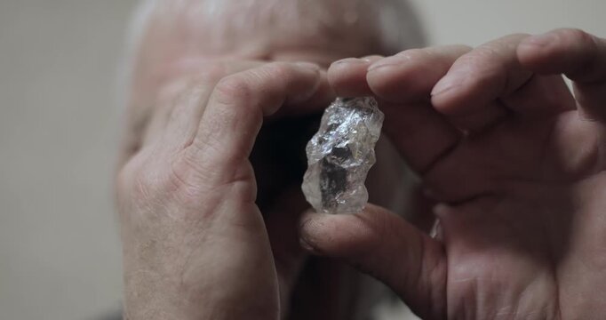 Tracking shot of man inspecting rough translucent gemstone between hands indoors, closeup revealing textured crystal.