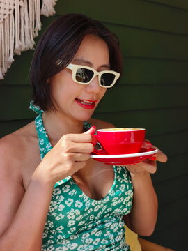 A stylish woman enjoys a cup of coffee while wearing white sunglasses and a green floral dress
