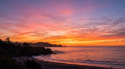 Obraz premium A vibrant sunset over the ocean with a rocky shoreline and distant mountains in the background.