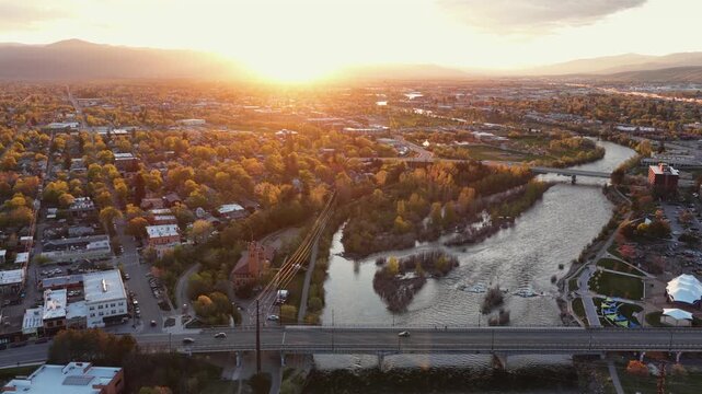 Missoula glowing in intense golden sunset light, Clark Fork River winding through autumn tree canopy, road bridge, riverside park and mountain range silhouette. Montana, USA. Aerial lateral wide shot