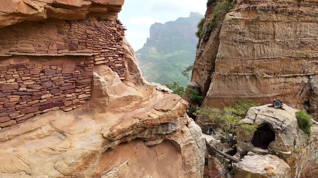 ETHIOPIA: Cliffside view of an ancient stone monastery built into sandstone rock faces in Tigray's Gheralta mountains, capturing a narrow ledge path and a scenic valley background.