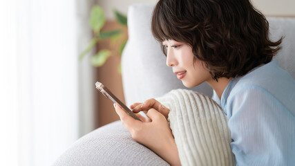 ソファで寝転びながらスマートフォンを使う女性, Woman Using Smartphone While Relaxing on a Sofa © Trickster*