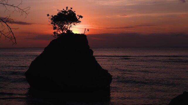 Padang Padang Beach (Pantai Labuan Sait) at sunset.  Island rock with tree in the foreground, with deep reds and purples of the setting sun in the background. The waves of the ocean can also be seen. 