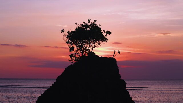 Padang Padang Beach (Pantai Labuan Sait) at sunset.  Island rock with tree in the foreground, with deep reds and purples of the setting sun in the background. The waves of the ocean can also be seen. 