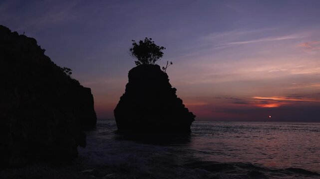 Padang Padang Beach (Pantai Labuan Sait) at sunset.  Island rock with tree in the foreground, with deep reds and purples of the setting sun in the background. The waves of the ocean can also be seen. 