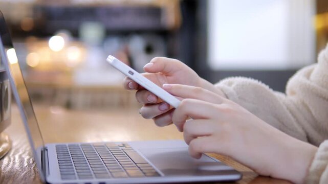Person uses phone while laptop sits open nearby. Hands hold phone steady. Background shows blurred cafe setting. Warm lighting highlights wooden table surface. Focus remains on hands and devices