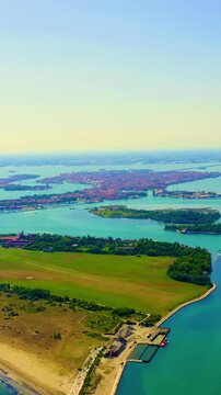 Vertical video. Venice, Italy. Aerial panoramic sweep over the Venetian Lagoon, scattered islands and terracotta rooftops of the historic city set against turquoise waters under clear summer sky. Dro