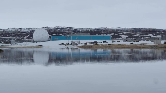 Ground-level camera shot of a large white radome dome and blue industrial building perfectly reflected in a calm pool of water during spring thaw in Iqaluit.
