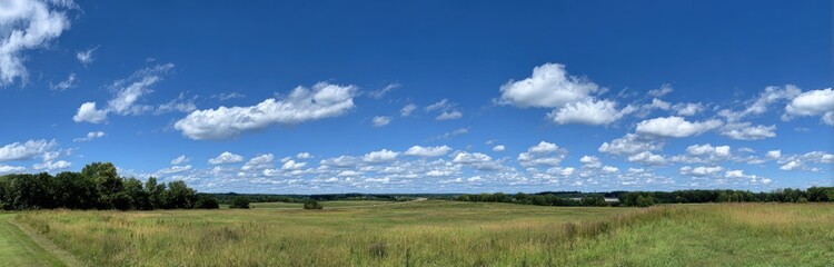 Obraz premium View of an expansive blue sky filled with fluffy white clouds over a lush green landscape with grassy fields and distant trees