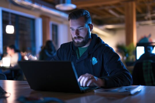 A focused man with a beard works on a laptop in a dimly lit modern office with exposed beams, surrounded by blurred coworkers.
