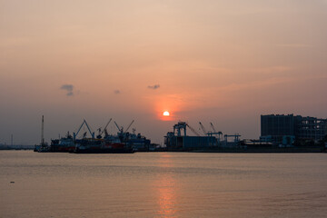 Obraz premium Sunset glows over calm harbor waters. Silhouetted cranes stand tall on dockside. Buildings and ships frame the fading light. Golden reflections ripple across the surface