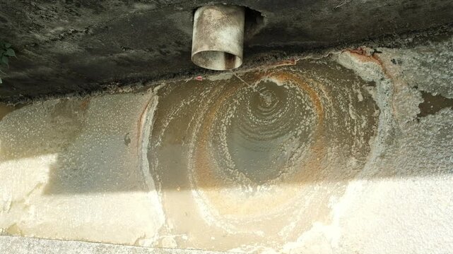 Polluted drain with oily scum and wastewater trickling from pipe.
