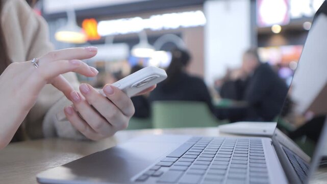 Person holds phone, taps screen. Laptop rests nearby, open on table. Background shows blurred public space with people. Focused, calm user engages digital device. Ideal for tech, communication