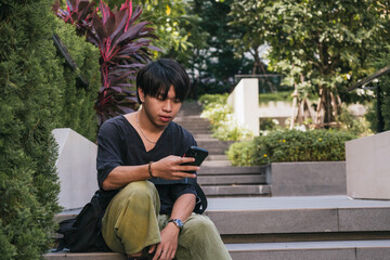 Fototapeta premium Young Asian man sitting relaxed on garden stairs, concentrating on his mobile phone in an outdoor setting