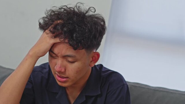 Young Asian man sitting on sofa shaking head suffering from severe vertigo