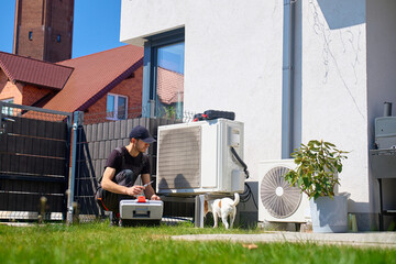Specialist servicing outdoor heat pump unit while a small dog stands near the equipment. Person inspecting heat pump on lawn near house on sunny day. HVAC service concept © Lazy_Bear