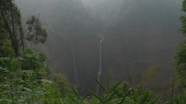Camera moves sideways to the right along a viewpoint, keeping Tad Fane&rsquo;s twin waterfalls in view behind jungle foliage in a hazy valley in Laos.