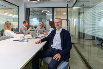 Fototapeta premium Confident stylish european man sitting at workplace, executive leader manager smiling, colleagues on the background