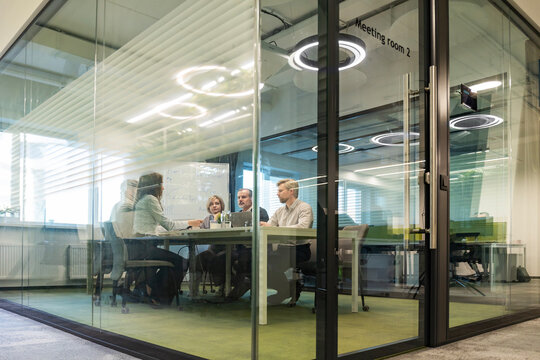 Group of business people standing together and discussing their work and projects, having a team meeting in an office, view from glass wall