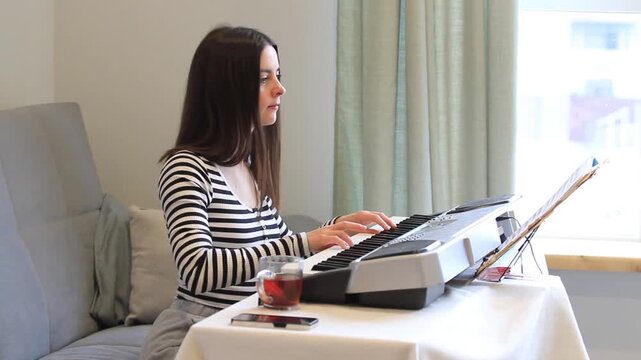 Musician woman playing electronic piano at home. Focused pianist practicing musical sheet. Online music lesson concept for composer skill development and hobby