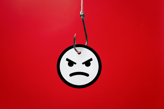 A metallic fishhook piercing an angry face emoji on a red background. Rage bait, social media trolling, online manipulation, clickbait traps, and deliberate digital provocation