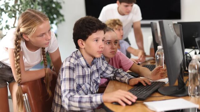Teenage boy sitting at table and using computer during computer science lesson. Teacher and classmates standing and looking at monitor