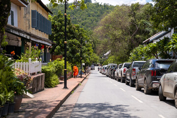 Naklejka premium Buddhist Monk Walking Along Peaceful Heritage Street with Colonial Buildings, Parked Cars, Green Hills, and Tropical Trees in Southeast Asia