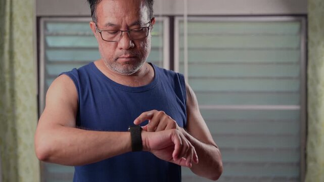 Asian man wearing glasses stands indoor, tapping smartwatch, checking health stats, tracking daily fitness progress, viewing workout data, planning active life routine, keeping fit, managing personal.