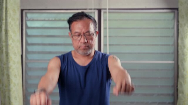 Determined middle-aged Asian man wearing eyeglasses, dark sleeveless shirt stretches both arms forward, clenching fists, performing home fitness routine, serious expression visible near glass window.