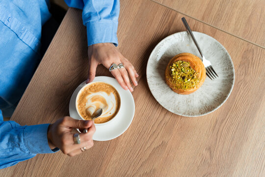 High angle view of a woman's hands stirring a cappuccino next to a sweet pistachio pastry on a wooden table