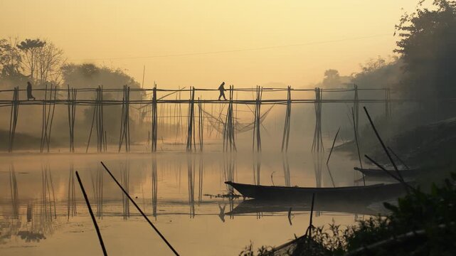 View of people walking across a traditional bamboo bridge over a misty river with fishing nets at sunrise Bogura, Rajshahi Division, Bangladesh.