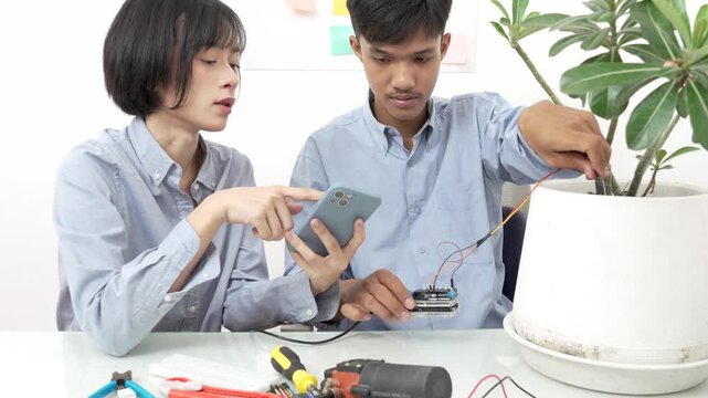 Young Asian woman and man using microcontroller and soil moisture sensors to measure soil moisture in a potted plant.