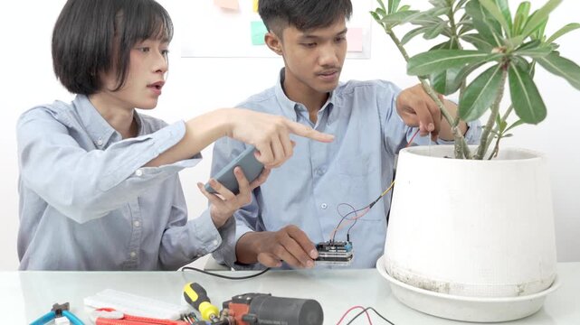 Young Asian woman and man using microcontroller and soil moisture sensors to measure soil moisture in a potted plant.